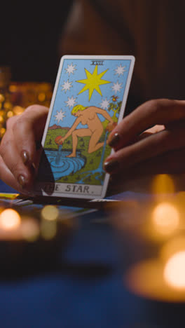 Vertical-Video-Close-Up-Of-Woman-Giving-Tarot-Card-Reading-On-Candlelit-Table-Holding-The-World-And-The-Star-Cards